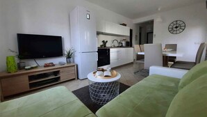 A modern living room with a flat-screen TV, a white refrigerator, a dining area with a table and chairs, and a kitchen area with cabinets and a sink.
