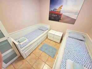 A small, compact room with two single beds, a small dresser, and a window with a beach scene.