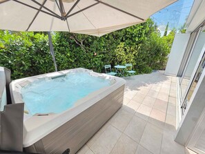 A hot tub under an umbrella, surrounded by greenery and a tiled patio.