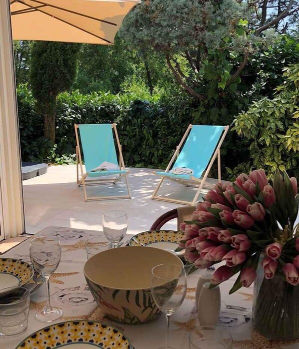 An outdoor dining area with a table set for two, surrounded by lush greenery and a large umbrella providing shade.