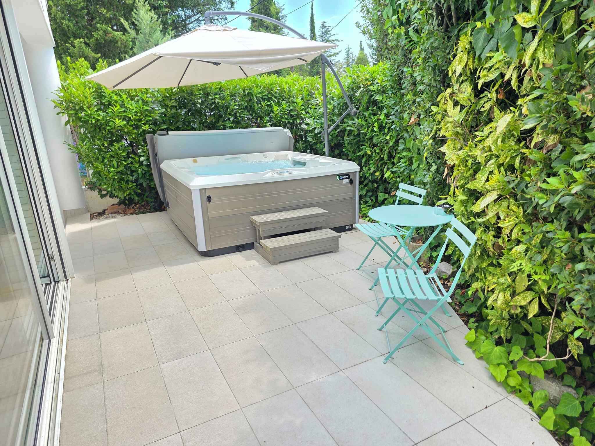 Ein Whirlpool mit beiger Abdeckung, ein Tisch und Stühle, umgeben von viel Grün.
