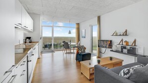 Una sala moderna con sofá, mesa de centro y televisor. Cuenta con una zona de cocina con armarios y una mesa de comedor con sillas. La habitación tiene ventanas amplias y vistas al exterior.