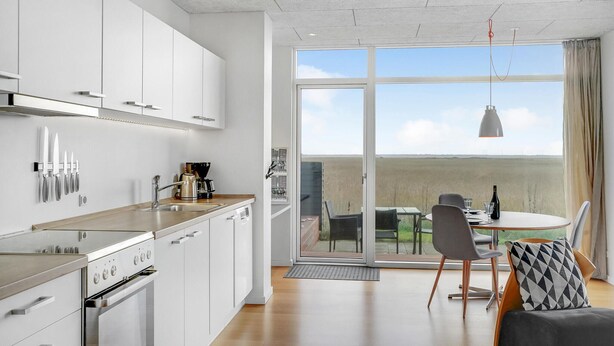 Una cocina moderna con gabinetes blancos y electrodomésticos de acero inoxidable, un comedor con mesa y sillas, y un amplio ventanal corredera que ofrece vistas al campo.