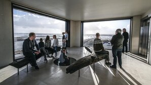 Un interior moderno con amplios ventanales que dan a una playa, un banco de madera y un grupo de personas reunidas.