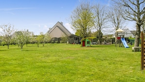 Ein grünes Feld mit einem kleinen, rot-gelben Spielhäuschen, einer blauen Rutsche und einem großen Gebäude im Hintergrund.