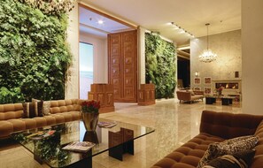 A spacious lobby with a large sofa, a glass coffee table, and a fireplace. The walls are adorned with lush green foliage, and there is a wooden door leading to another room. - Nice apartment in Binz with house sea view (Prora/Rügen)