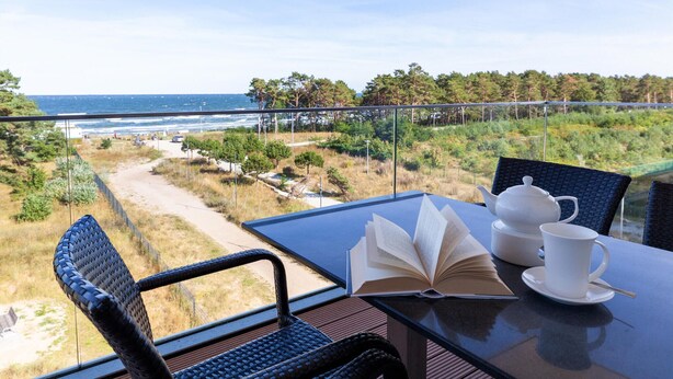 Et bord med en bog, tepotte og kop på en balkon med udsigt over strand og skov.