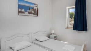 Ein Zimmer in Weiß mit einem Bett, einem Nachttisch, einer Lampe, einem Fenster mit Vorhang und einem Bild an der Wand.