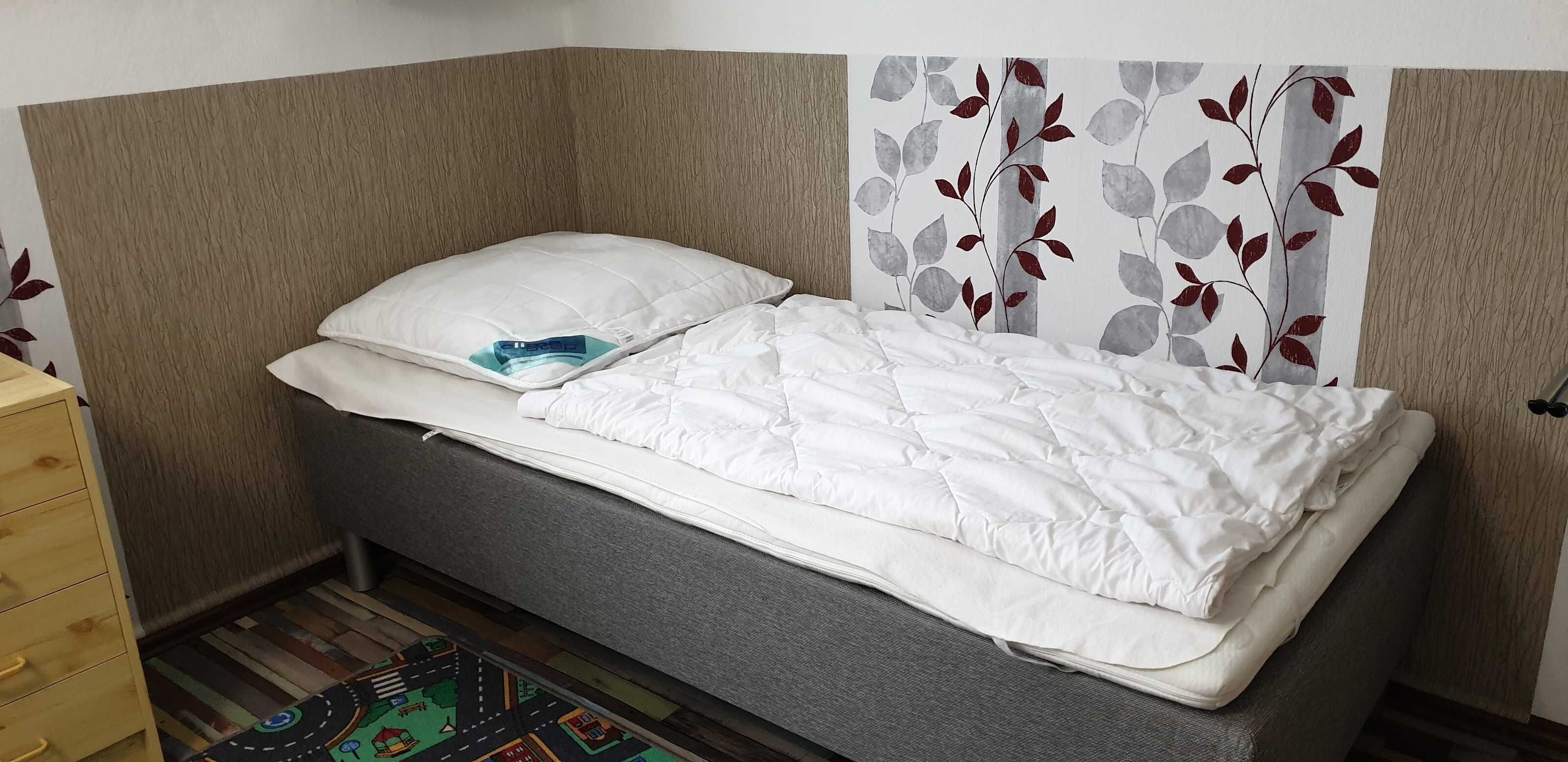 Un lit simple avec une couette et des oreillers blancs, une petite table de chevet et un mur orné d’un motif de feuilles.