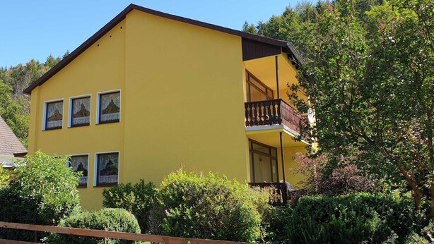 Edifício amarelo de dois andares com varanda, rodeado por vegetação.