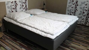 Uma cama com colchão e almofadas brancas, uma mesinha de cabeceira com abajur e uma parede estampada.