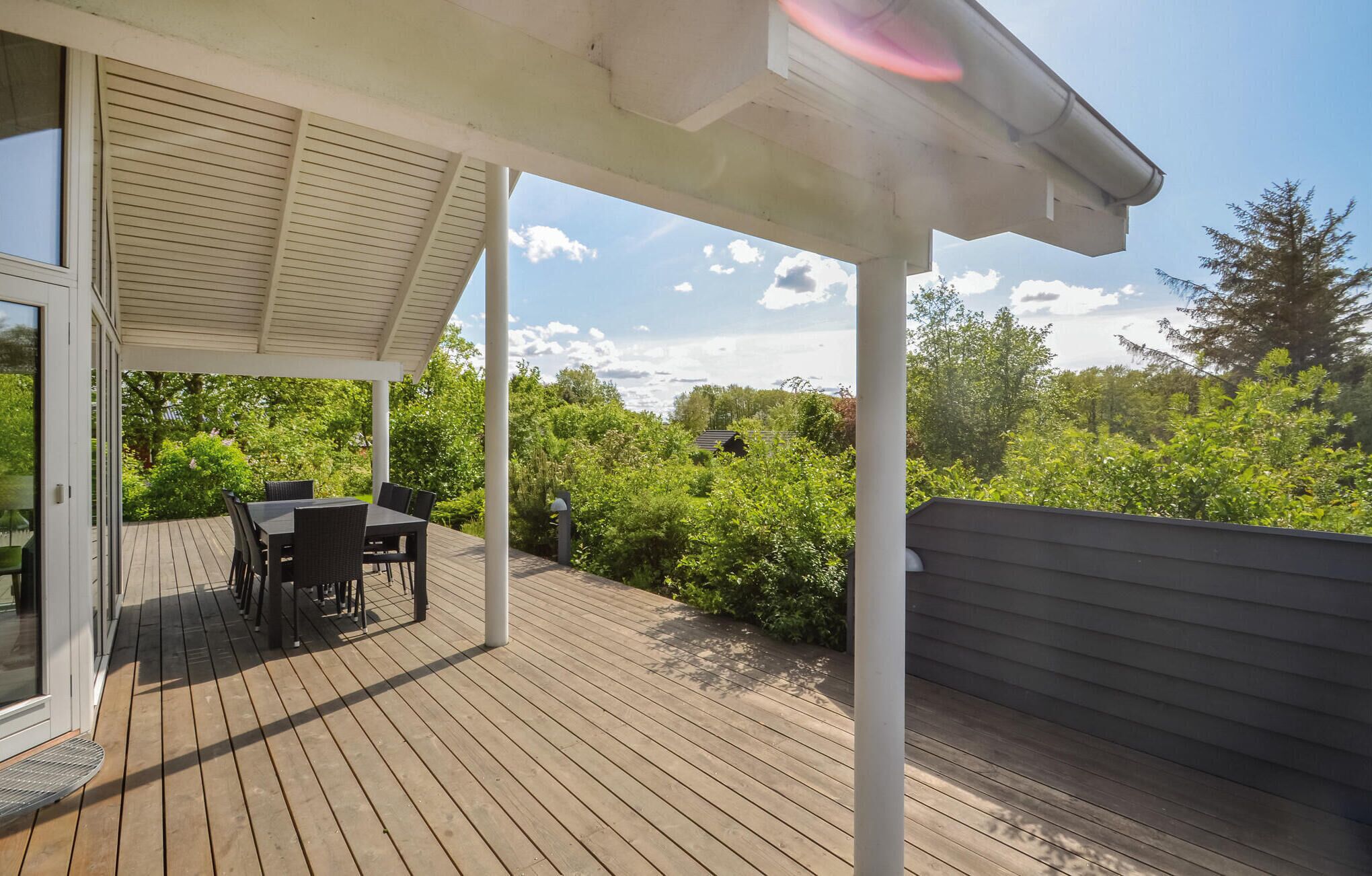 A wooden deck with a table and chairs, surrounded by greenery and a clear sky.