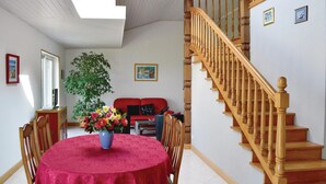 Un comedor con una mesa redonda cubierta por un mantel rojo, rodeada de sillas de madera. Una planta grande en maceta está en el centro, y un sofá rojo está apoyado contra la pared. Se ve una escalera de madera con barandilla a la derecha.