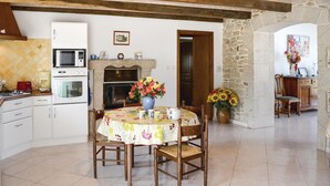 Cocina con armarios blancos, microondas, horno y comedor con una mesa para dos. Se observa una pared de piedra y una puerta de madera que da a otra habitación.