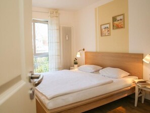 A bedroom with a wooden bed, white bedding, a bedside table with a lamp, and a window with curtains. - (102) 2-room apartment - lake view - apartment complex Baltic Sea view (Kühlungsborn)
