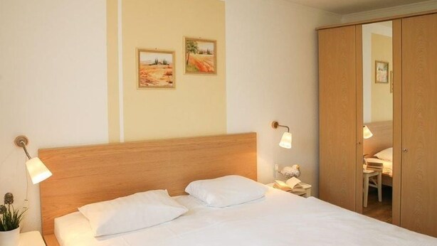 Una cama bien tendida con sábanas blancas, cabecera de madera y dos lámparas fijadas a la pared. Encima de la cama, dos cuadros enmarcados.