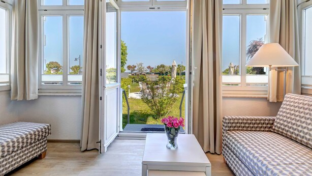 Ein Zimmer mit einer Couch, einem kleinen Tisch mit Blumenvase und Blick auf einen Garten durch ein großes Fenster.