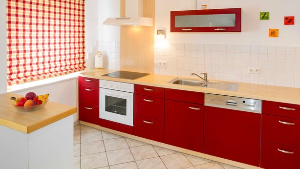A kitchen with red cabinets, a white stove, a dishwasher, and a window with a checkered blind. - FW I - Pension ANNA, Apartments & Comfort Rooms *** (Keuschen)