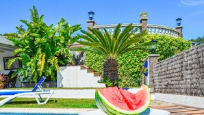 Une piscine entourée d’une terrasse en bois, d’une clôture blanche et d’une végétation luxuriante comprenant des bananiers et un palmier.