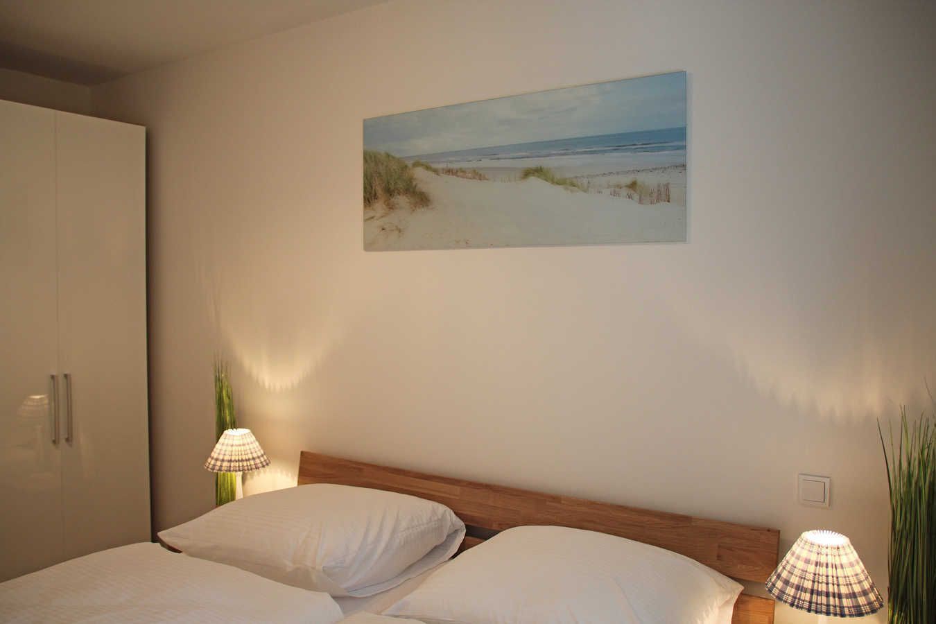 A bedroom with a bed, a wardrobe, a painting of a beach, and two lamps.