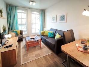 A modern living room with a sofa, coffee table, and a dining table. There are large windows with curtains, a TV, and a lamp. - Haus Meeresblick FeWo Strandkrabbe A 3.16 Ref. 159955 - FeWo Strandkrabbe A 3.16 (Baabe)