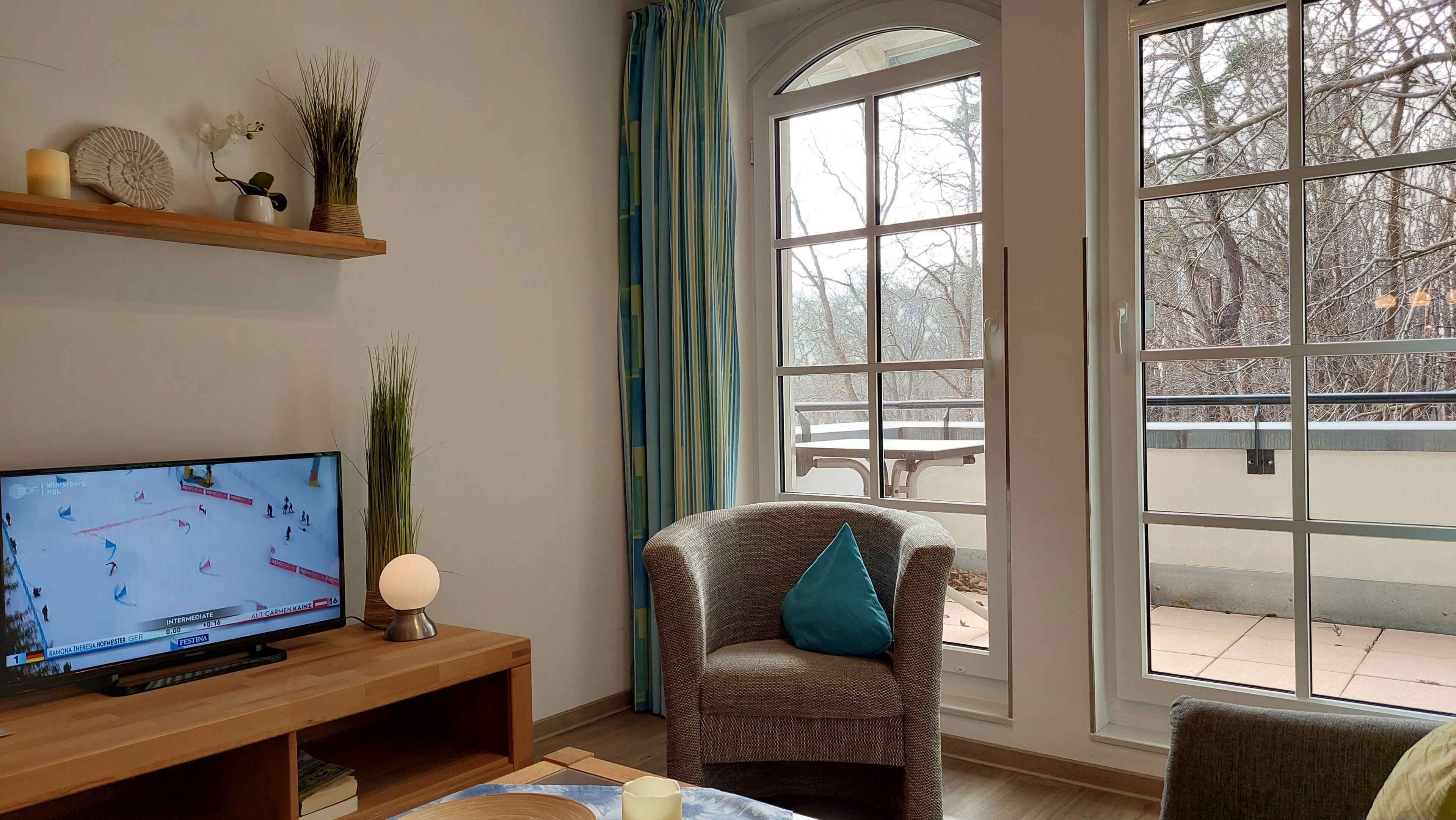 A living room with a TV, a chair with a pillow, a wooden shelf with decorative items, and a large window with a view of trees.