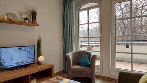 Una sala de estar con televisor, una silla con cojín, un estante de madera con objetos decorativos y un ventanal con vista a árboles.