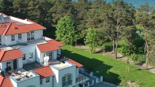 Un edificio de varios pisos con techo de tejas rojas, rodeado de vegetación y vistas al mar.