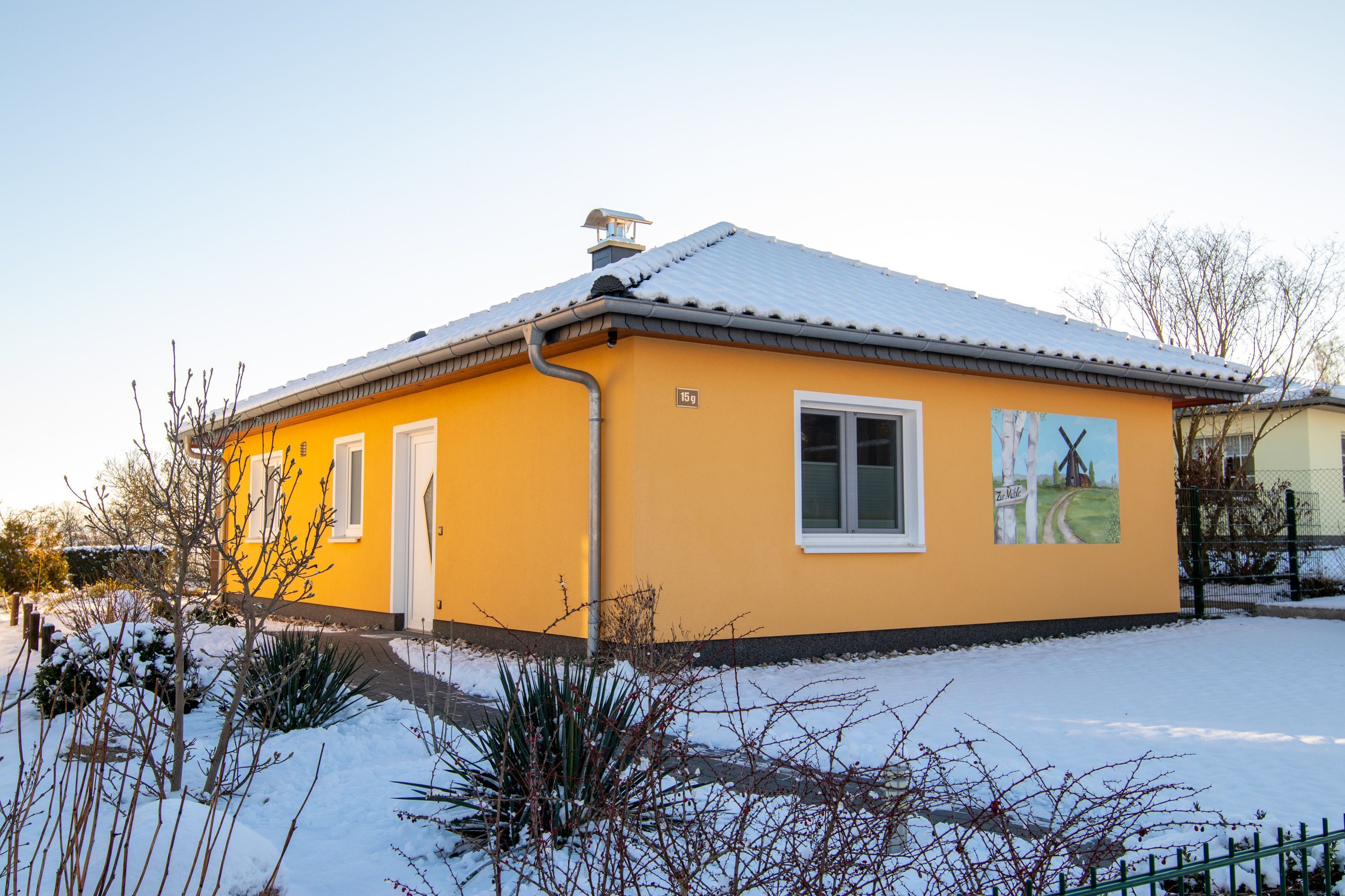 Un bâtiment jaune avec un toit enneigé, une cheminée et un petit jardin également couvert de neige.