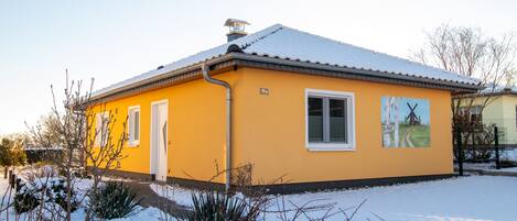 Un bâtiment jaune avec un toit enneigé, une cheminée et un petit jardin également couvert de neige.