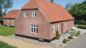 A two-story brick house with a steeply pitched red-tiled roof, a front porch, and a small garden with potted plants.