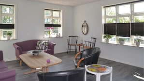 A modern living room with a purple sofa, a wooden coffee table, and a white side table with colorful glass tops. There are two black leather chairs, a small round table with a candle, and a mirror on the wall.