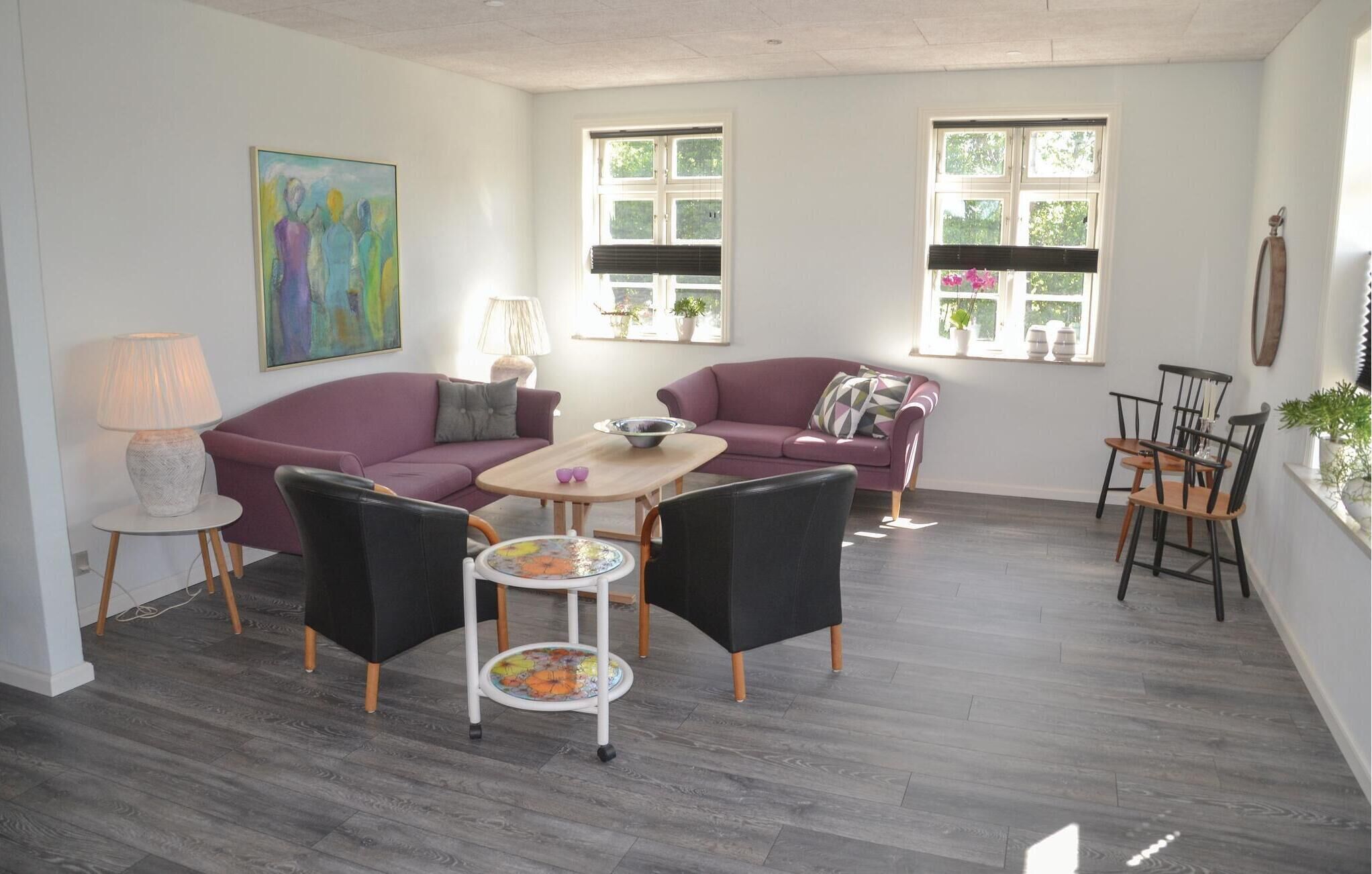 Une pièce avec deux canapés violets, une table ronde et une petite table à manger avec des chaises. Il y a deux fenêtres avec des stores, une lampe et un tableau au mur.
