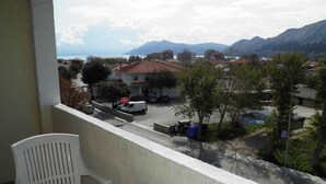 A balcony with a white table and chairs, overlooking a street with buildings, trees, and mountains in the background.