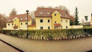 Keltainen rakennus, jossa on punatiilikatto, asfaltoitu ajotie ja etupuolella pensasaita.
