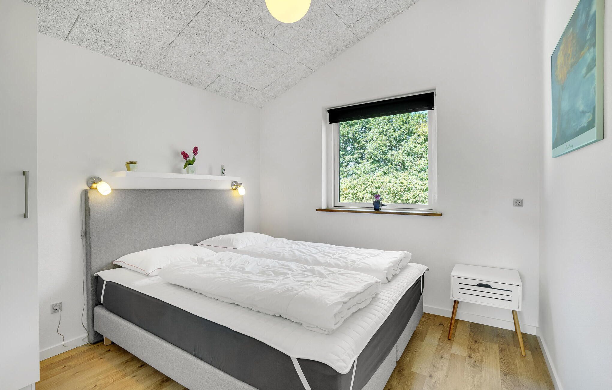 A bedroom with a bed, a window with a view of greenery, a bedside table, and a wall-mounted shelf with decorative items.