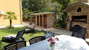 An outdoor patio with a table set for dining, surrounded by chairs, a small garden, and a stone building with a fireplace. - Holiday apartment with air conditioning (Malinska)