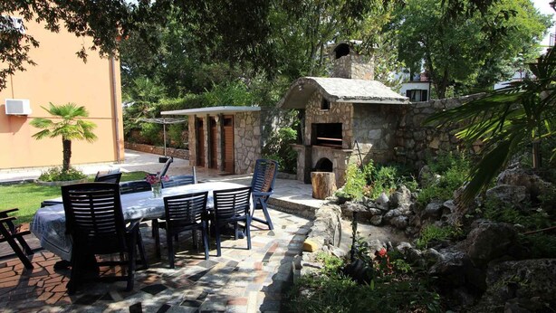 Eine Terrasse im Freien mit einem Stein-Barbecue und Sitzbereich, umgeben von Grünpflanzen und einer Steinmauer.
