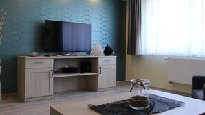 A modern living room with a flat-screen TV, wooden furniture, a glass vase, and a window with curtains. - Densee: Ferienwohnung Süderende - Apartment (Insel Hiddensee)