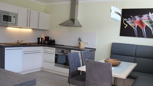 A modern kitchen with white cabinets, a built-in oven, and a microwave. There is a dining area with a table and chairs. - Densee: Ferienwohnung Süderende - Apartment (Insel Hiddensee)