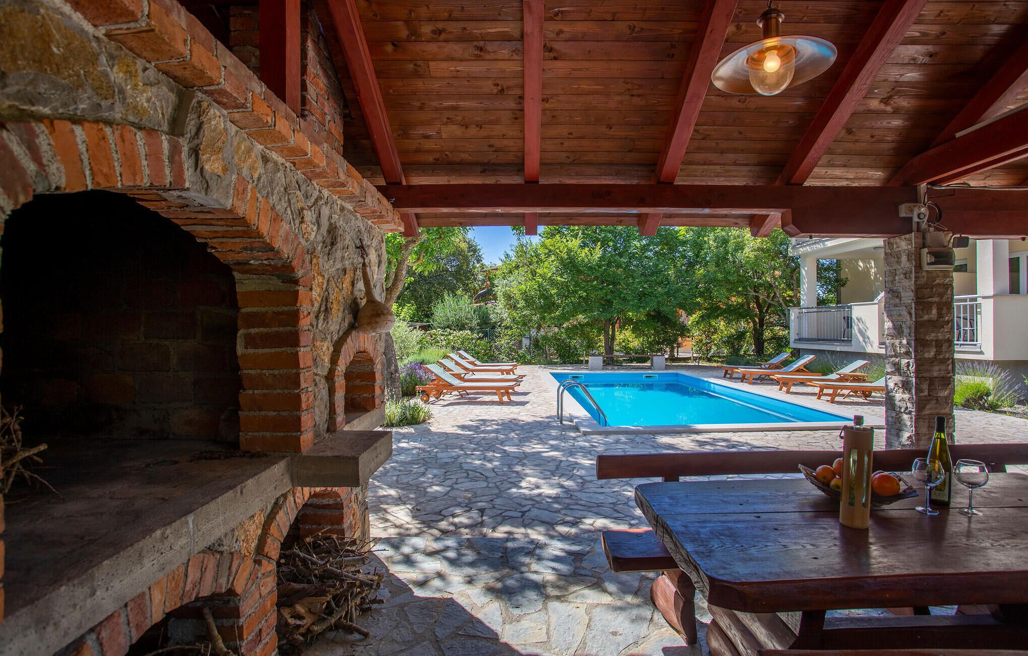 A pool area with a brick oven, lounge chairs, and a table with a bottle and glasses.