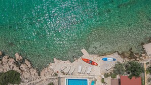 Vista aérea de una propiedad frente a la playa con piscina, sillones de descanso y kayaks.