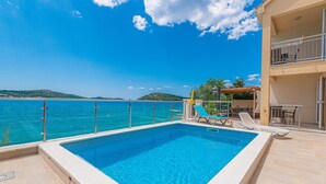 Una piscina rectangular con agua azul cristalina, rodeada por una terraza con baldosas. Se observa una pérgola de madera con una mesa y sillas al fondo.