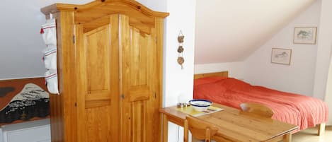 A bedroom with a bed, a wooden wardrobe, a dining table with a chair, and a wall-mounted shelf.