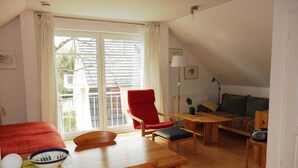 A room with a dining table, chairs, a red armchair, a sofa, a small table with a lamp, and a window with curtains. - Haus Bernstein 3 - Apartment Bernstein 3 (Graal-Müritz)