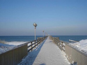 Une promenade en bois recouverte de neige, qui mène à l’océan, avec des garde-corps en bois et des lampadaires.