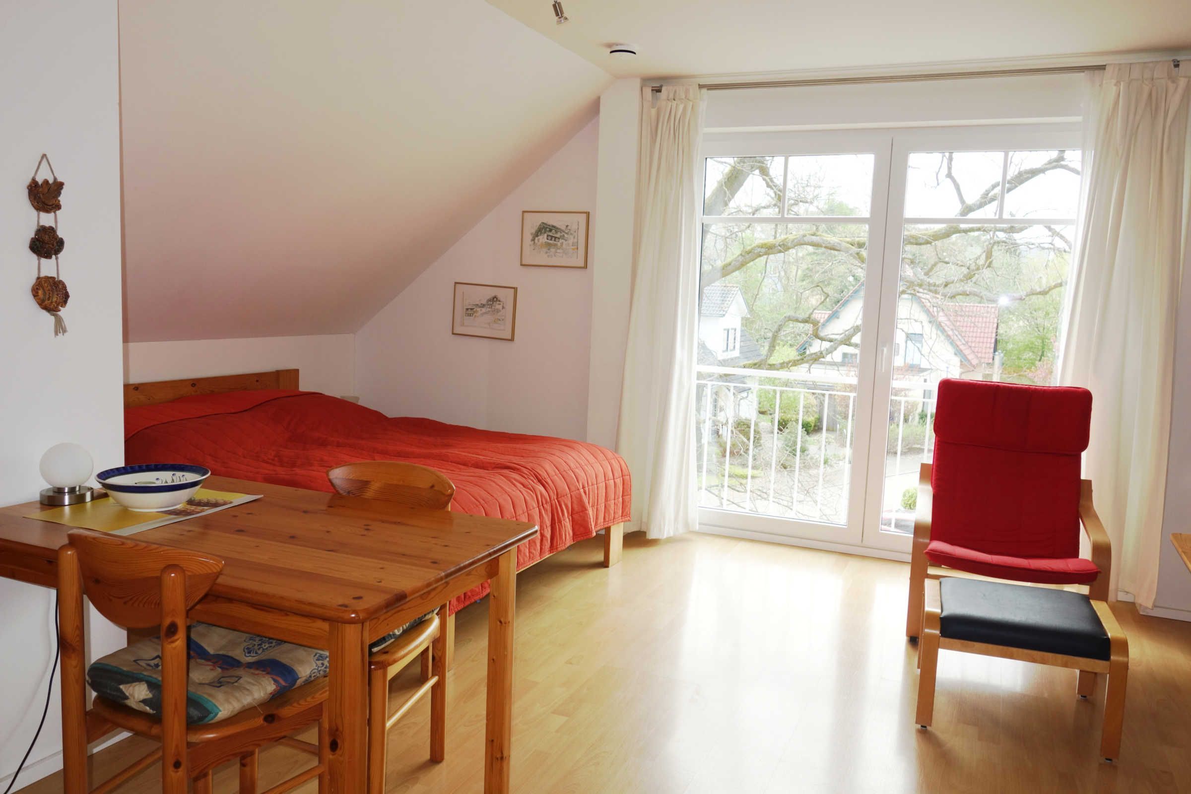 A bedroom with a bed, a table with chairs, a red chair, and a window with a view of houses.