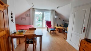 A room with a bed, a dining table, a red chair, a sofa, and a window with a view of greenery. - Haus Bernstein 3 - Apartment Bernstein 3 (Graal-Müritz)