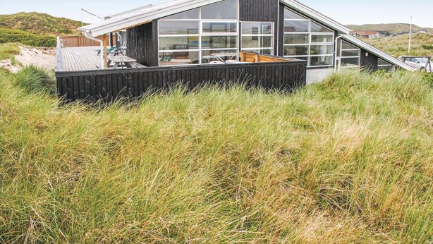 Ett modernt hus med en stor glasfasad, ett trädäck och en grönskande trädgård.