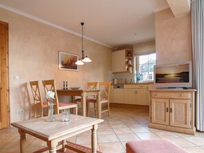 Ein Essbereich mit Holztisch und Stühlen, einem Gemälde an der Wand sowie einer Küche mit Schränken und einem Fernseher.